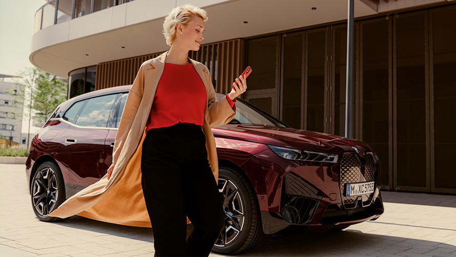 My BMW App My BMW App BMW iX i20 Woman walking in front of BMW vehicle with smartphone in hand